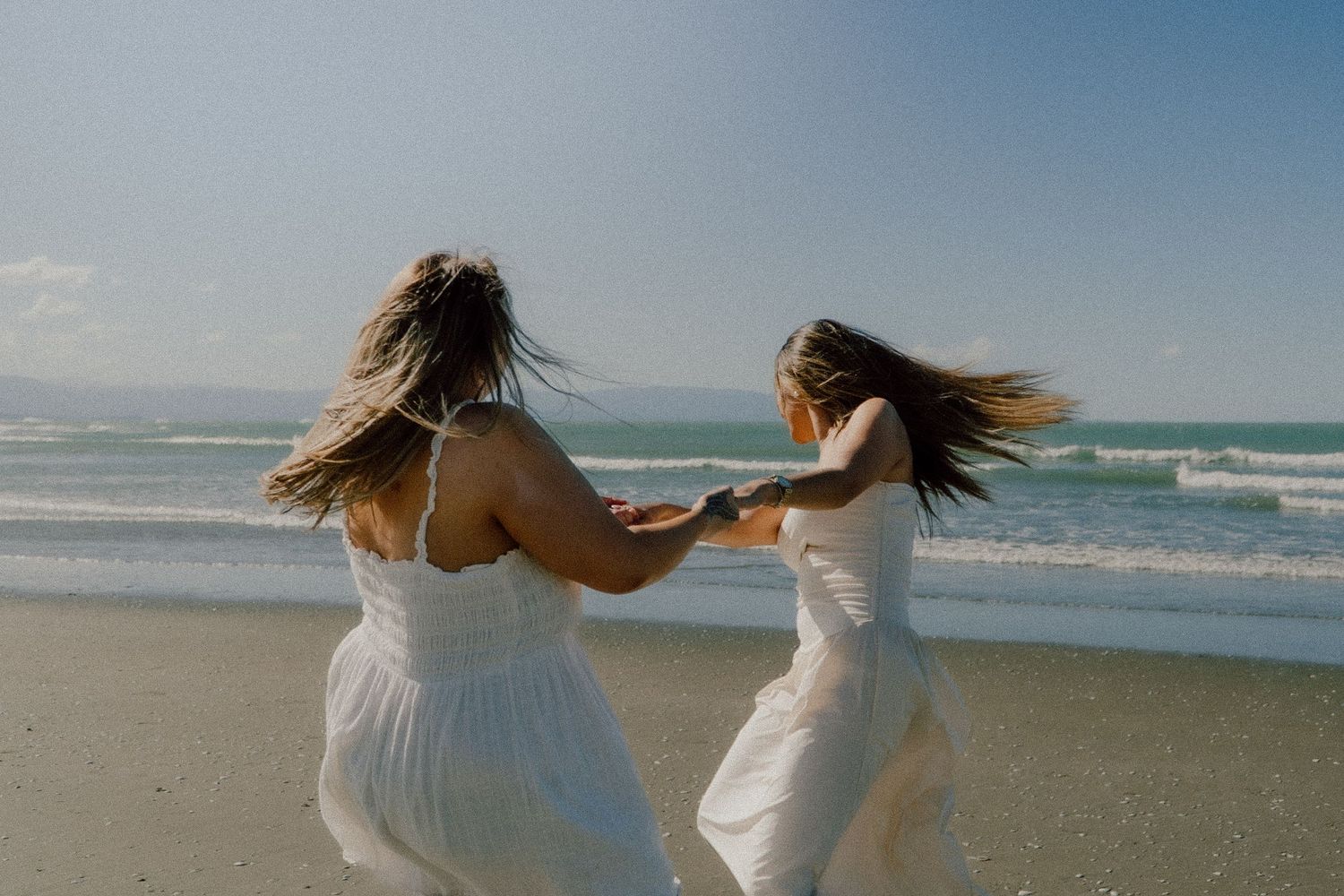Two friends pose together on a sunny beach with ocean waves in the background.
