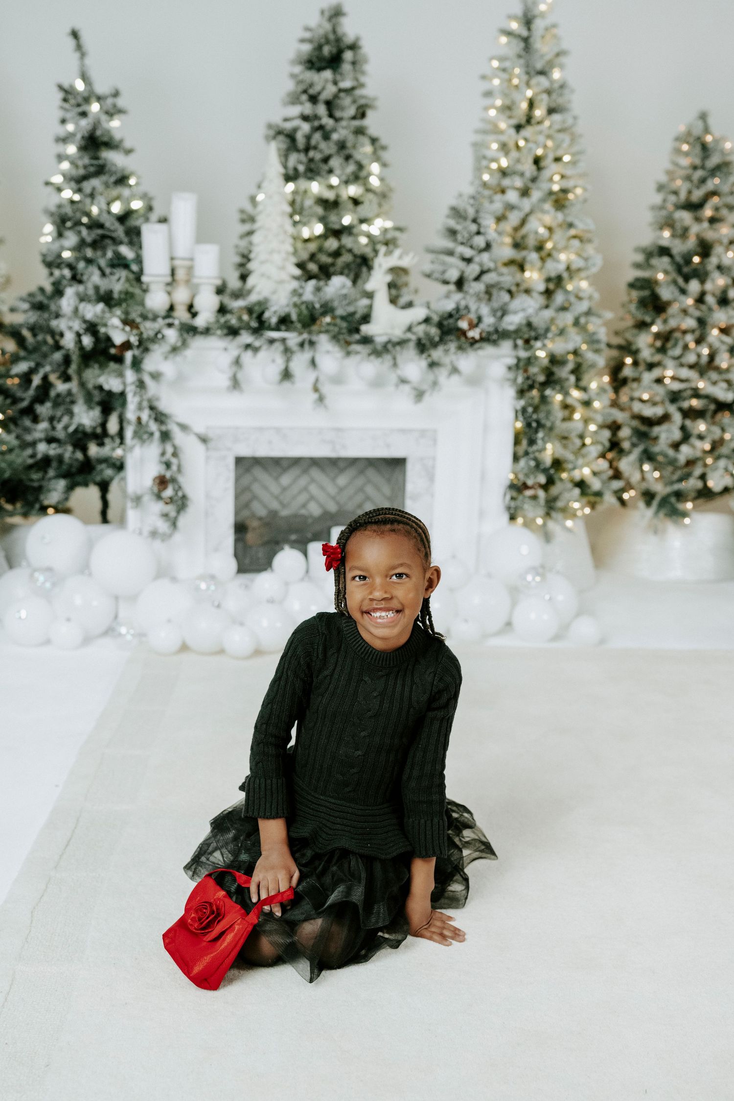 Child in black sweater poses against white winter holiday backdrop with decorated Christmas trees and twinkling lights.