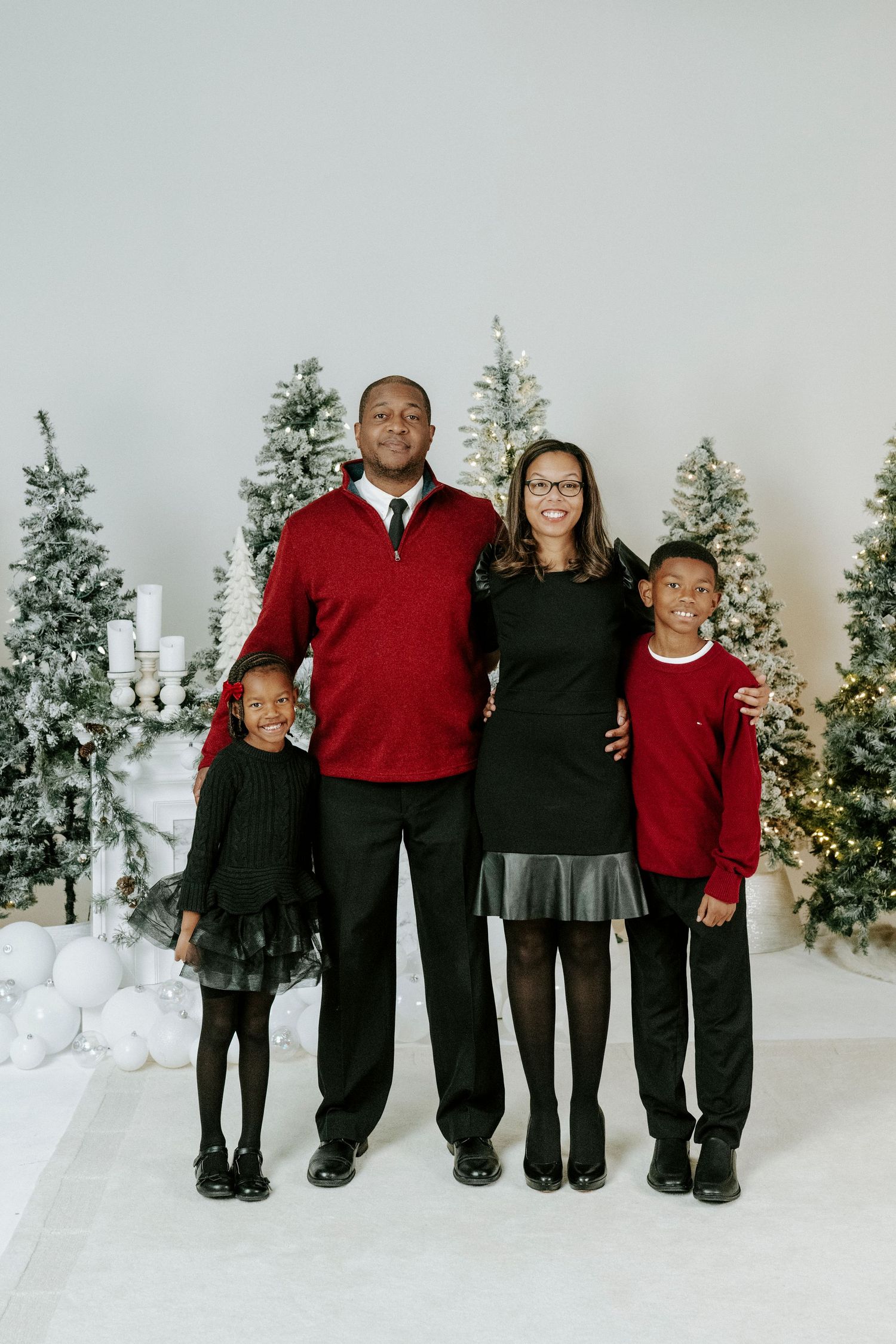 A family group poses together in coordinating red and black outfits against a backdrop of lit Christmas trees.