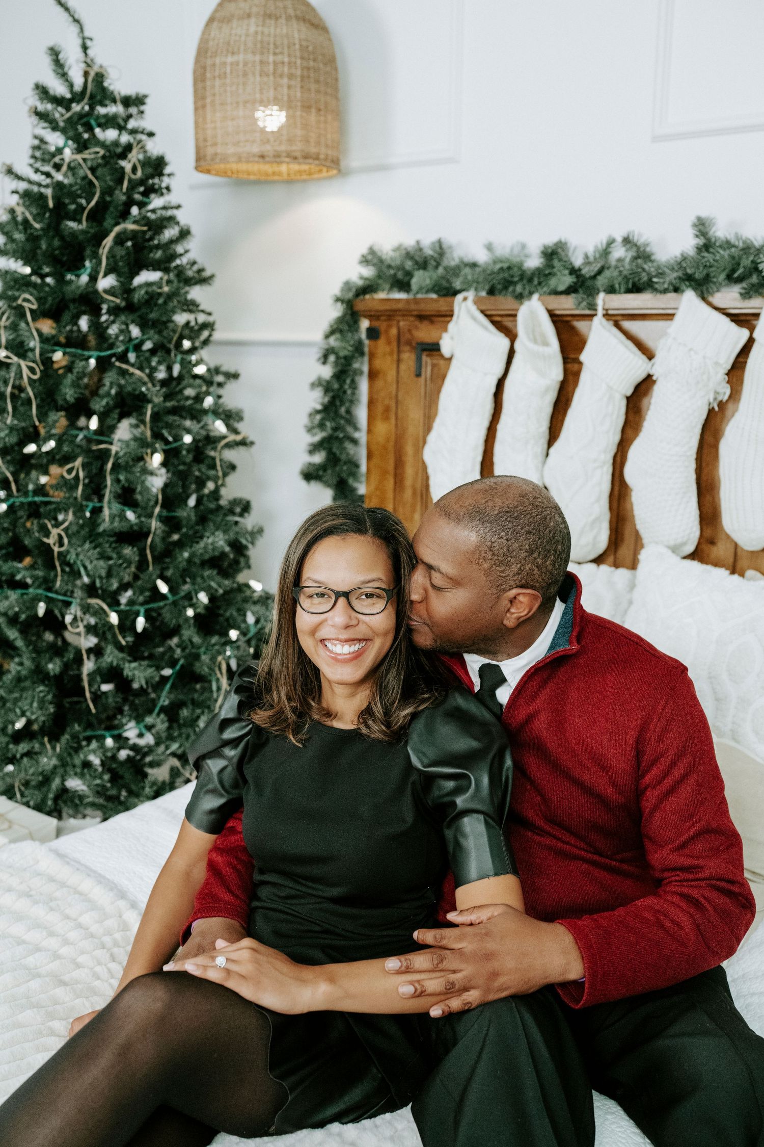 A couple in black and burgundy outfits share intimate moments in front of a Christmas tree and stockings.