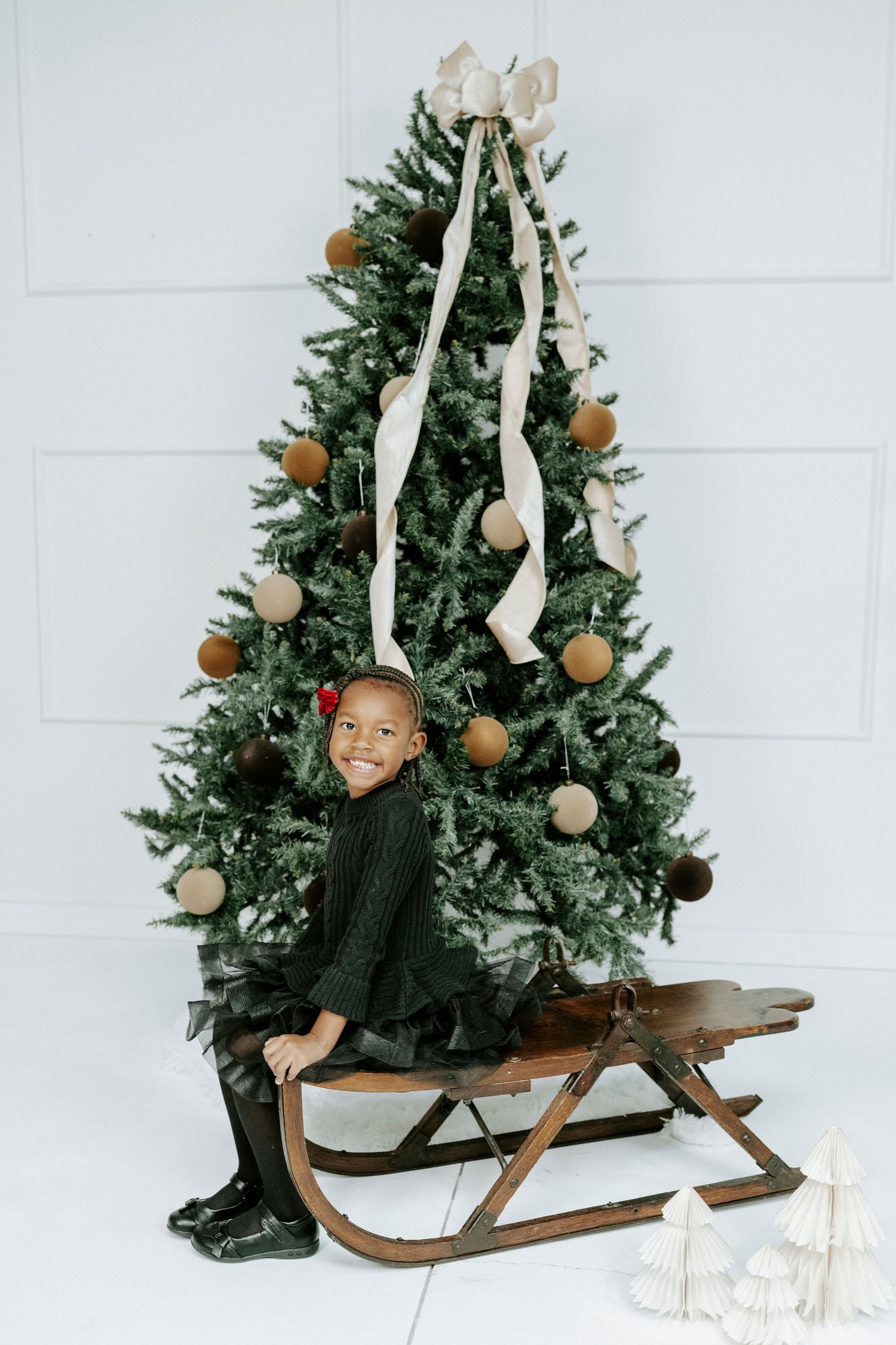 A Christmas scene with a decorated evergreen tree and vintage wooden sled against a white backdrop.