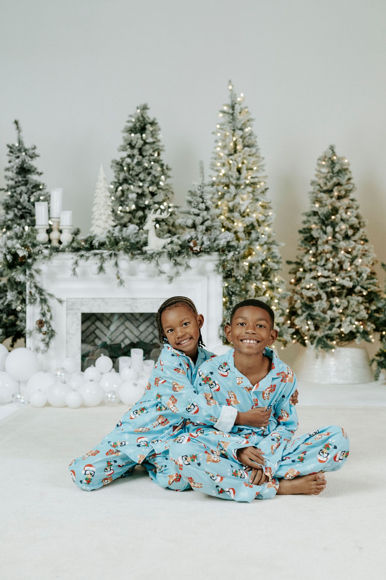 Two children in matching blue Christmas pajamas laying and playing on white floor with decorated trees in background.