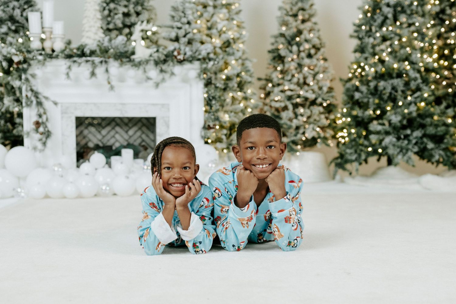 Two children in matching blue floral pajamas lie on white floor with decorated Christmas trees in background.