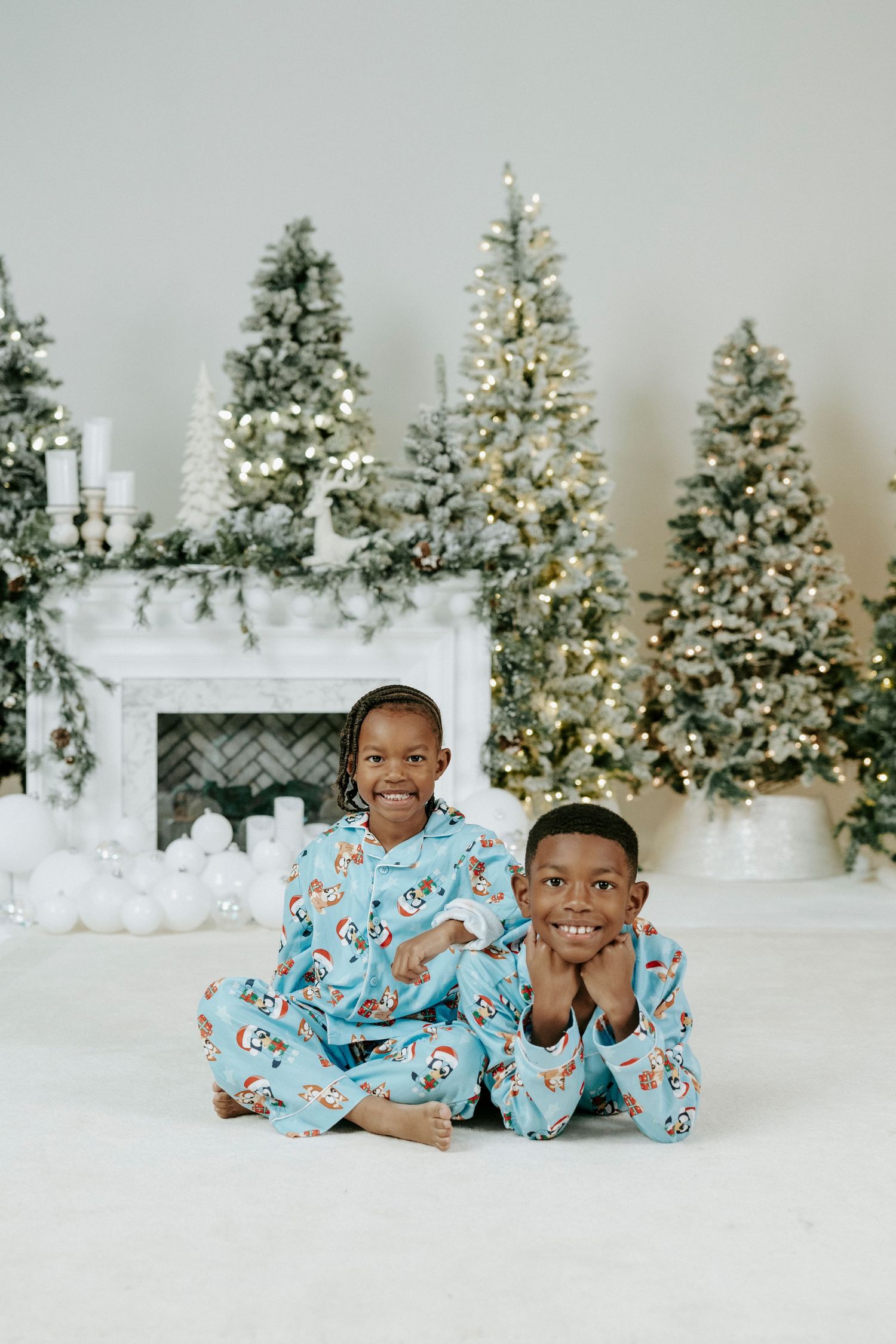 Two children in matching blue pajamas lay on white floor with decorated Christmas trees in the background.
