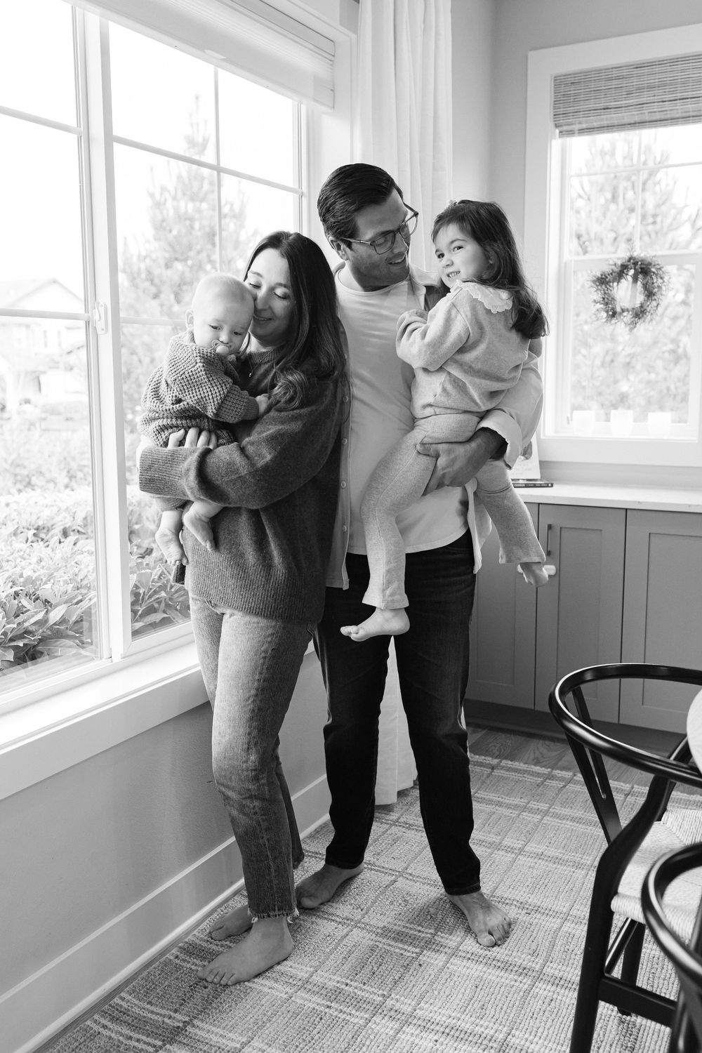 A series of black and white photos showing tender family moments by a bright window in a home interior.