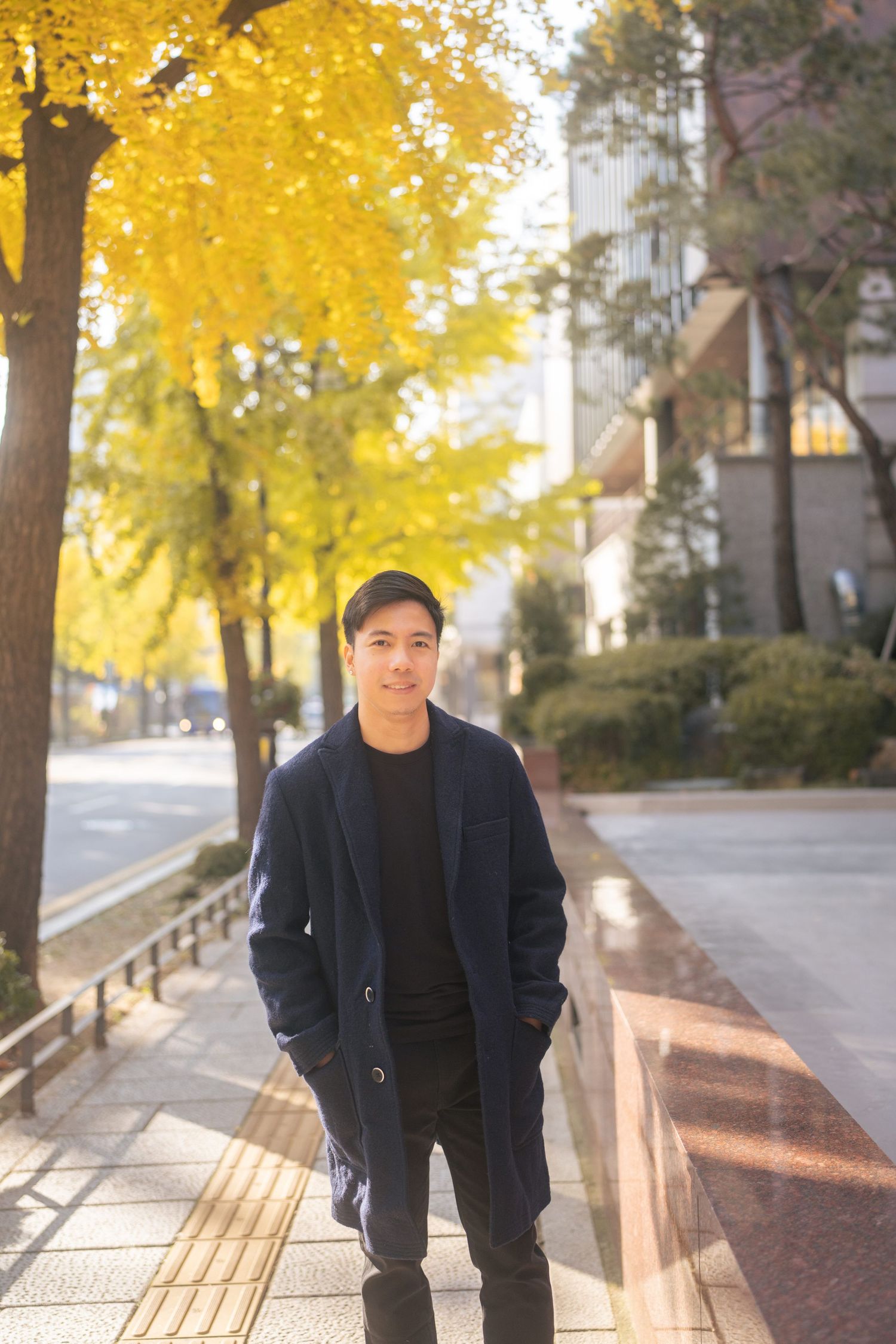 Person in black coat stands on wooden bench under vibrant yellow autumn trees in urban setting.