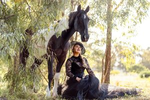 A dark horse stands beside a person in black clothing sitting on the ground under willow trees in natural sunlight photoshoot in Sydney