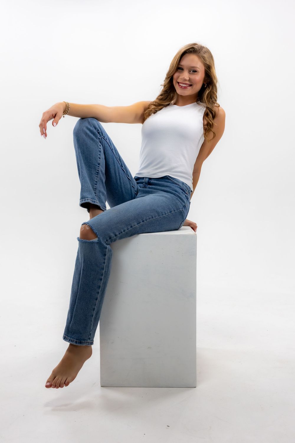 Casual portrait of someone in blue jeans and white tank top sitting barefoot on a gray cube against white background.