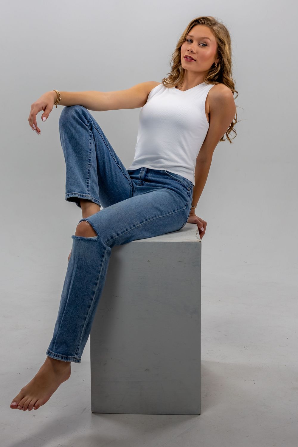 A person in blue jeans and white tank top poses on a gray cube against a light background in a fashion photo series.