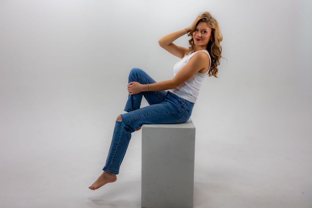 A figure in blue jeans sits casually on a white cube against a light gray studio backdrop.