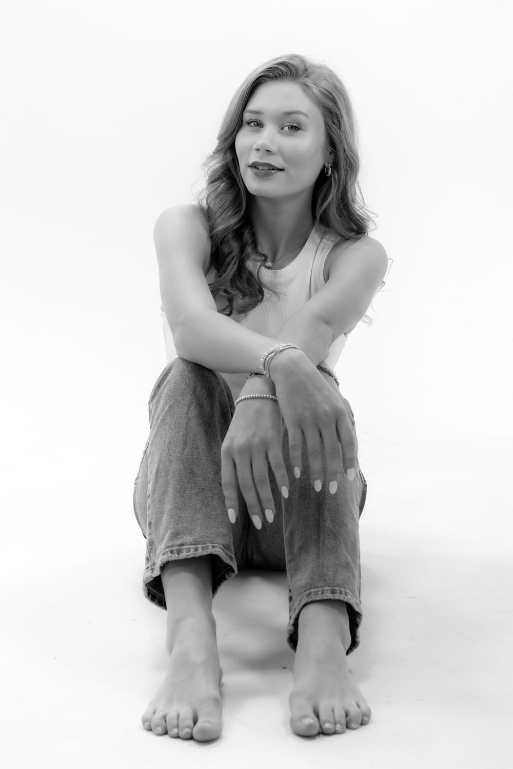 Black and white portrait of someone sitting casually in jeans and tank top against white background.
