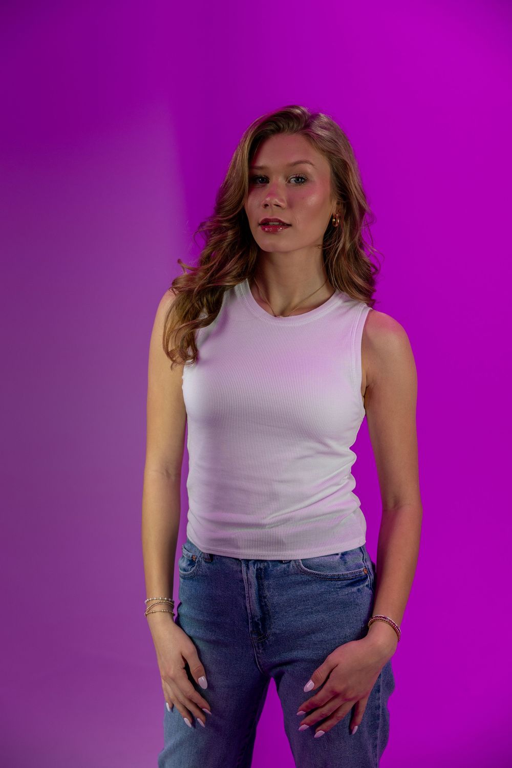 Person wearing white sleeveless top and blue jeans poses against vibrant purple background.