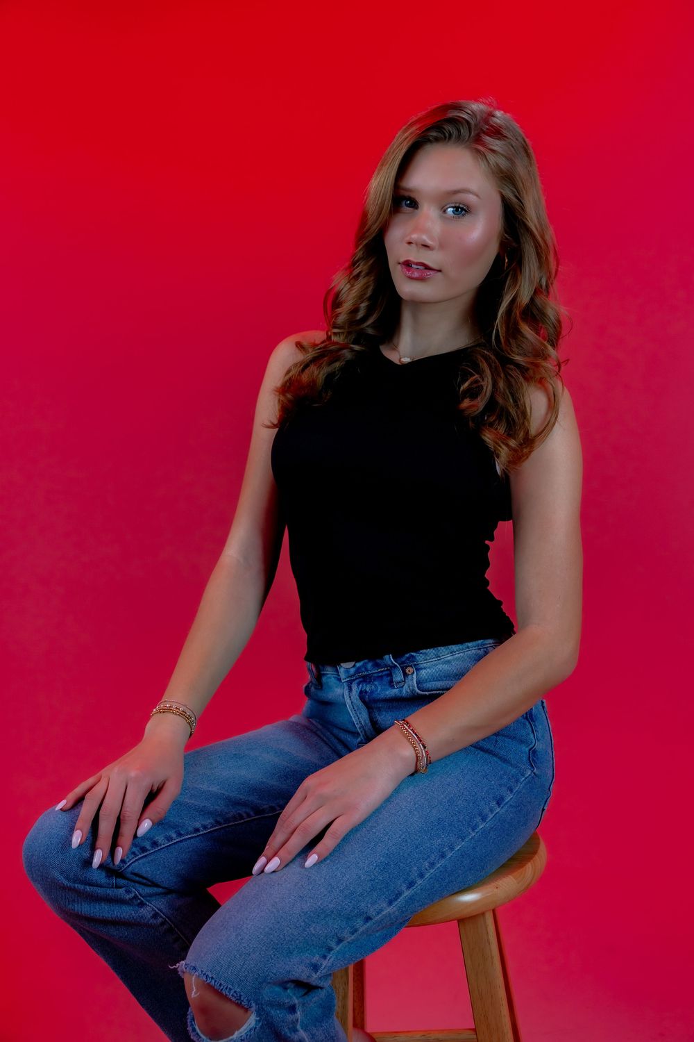 Fashion portrait of a model in black tank top and blue jeans posing against a vibrant red backdrop.