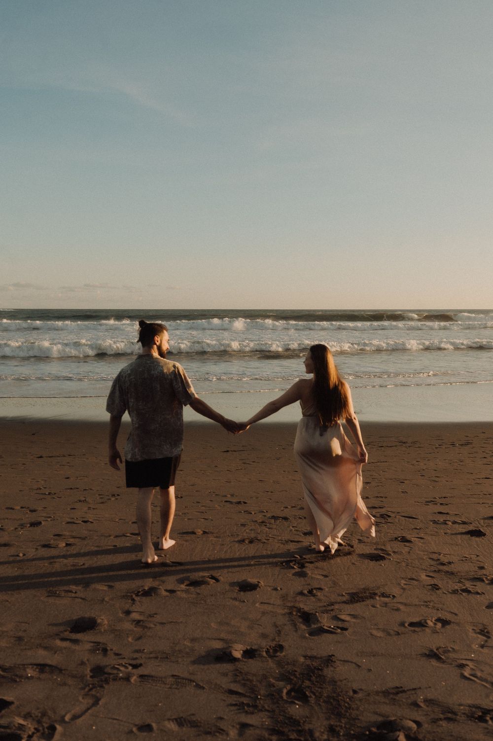 A couple walks together on a sandy beach at sunset with waves crashing in the background.