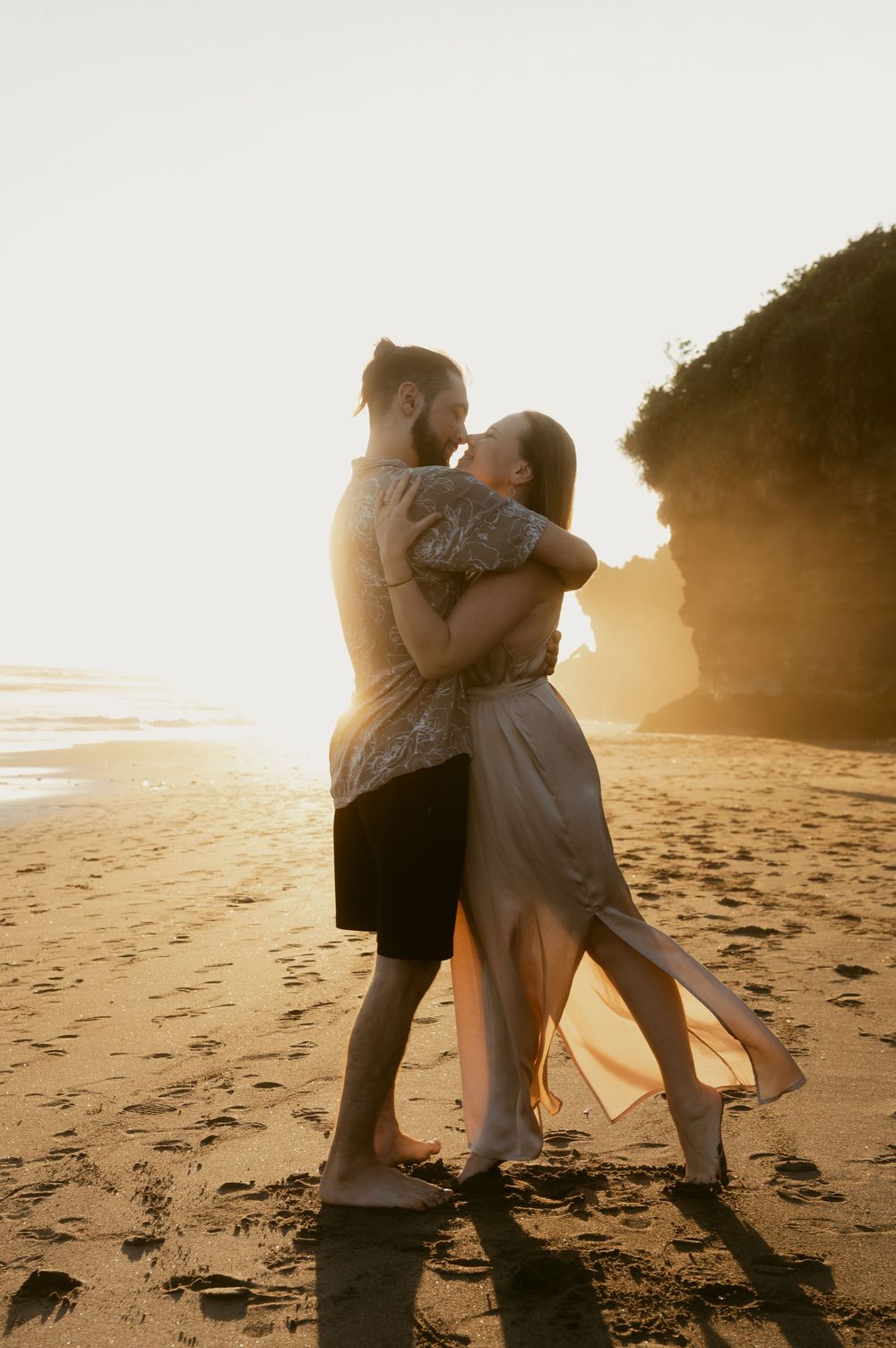 Romantic couple embracing on a sunlit beach during golden hour with cliffs in the background.