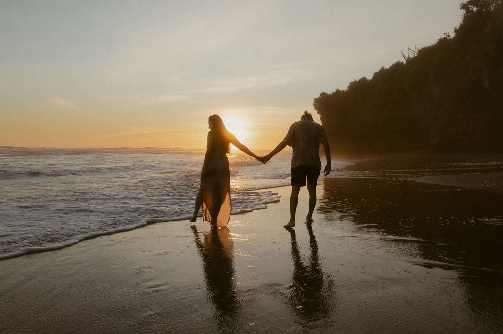 Two figures hold hands while strolling along a scenic beach at sunset with cliffs in the background.