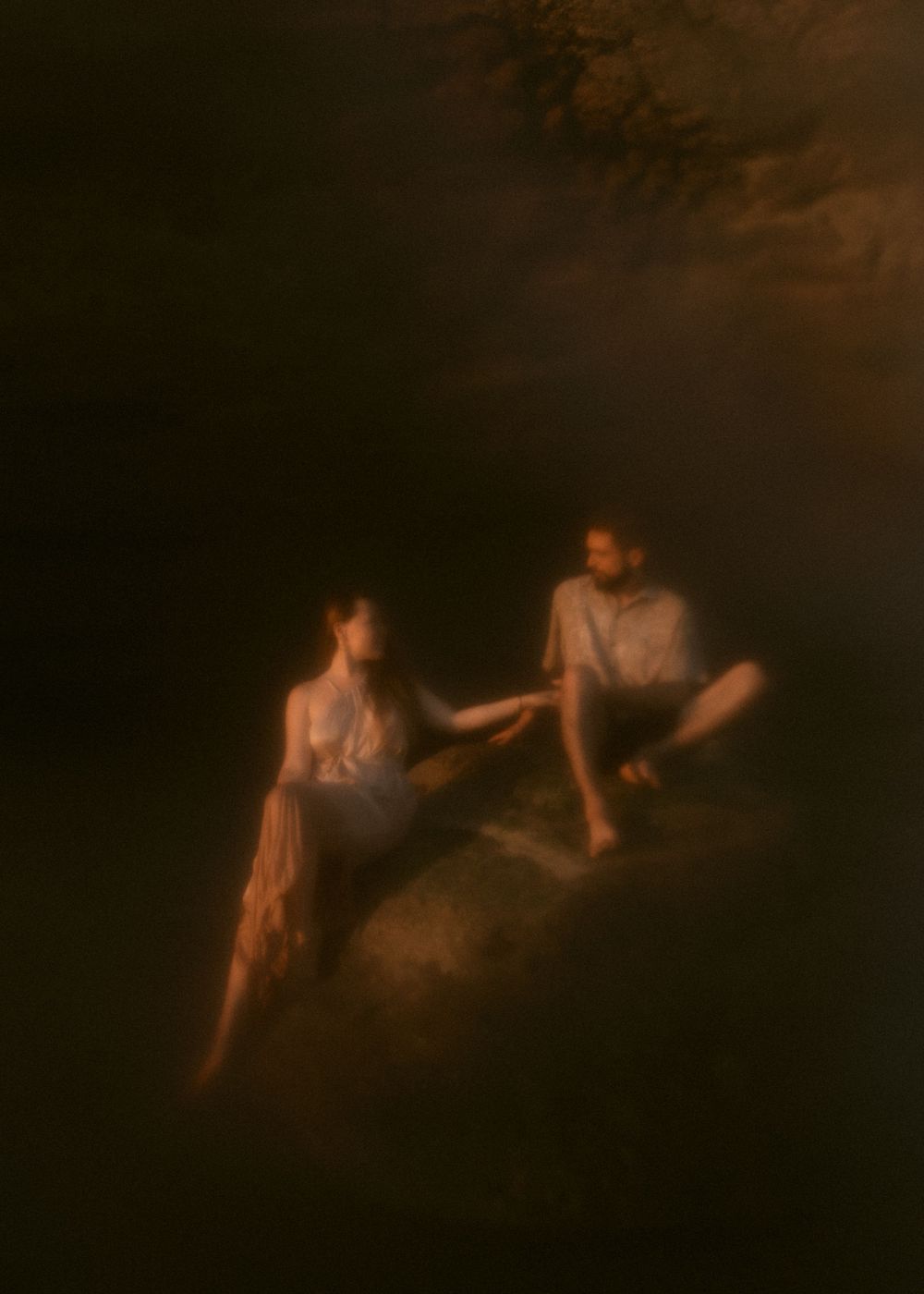 Two people in casual attire sit together on a dark beach during golden hour.