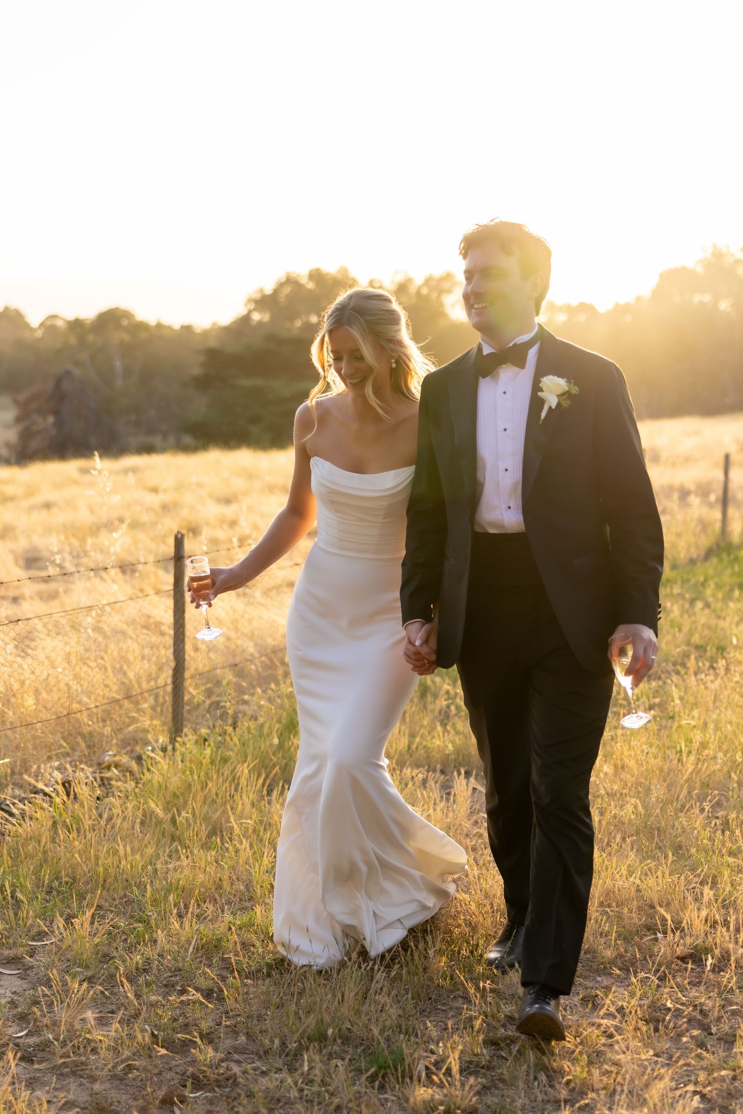 A bride and groom walk hand in hand through a sunlit meadow at golden hour creating a romantic silhouette.