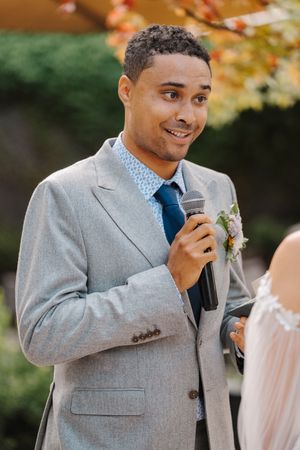 A groom in a gray suit and blue tie delivers an emotional wedding speech outdoors during an autumn ceremony.