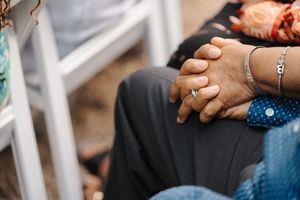 Close-up view of clasped hands wearing wedding rings and a bracelet, sitting on dark and blue denim.