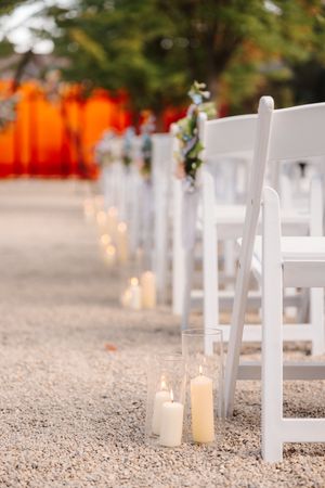 White folding chairs line a gravel aisle with glowing candles creating a romantic outdoor wedding ceremony setup.