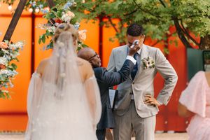 A couple poses with their cat during their outdoor wedding ceremony against a vibrant orange wall.