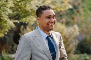 A person in a gray suit and blue tie smiles while standing outdoors in an autumn setting.