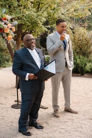 A joyful moment captured at an outdoor wedding with groomsmen in black and tan suits sharing laughter on a garden path.
