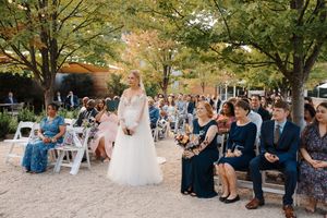 An outdoor wedding ceremony in a garden setting with guests seated on white chairs beneath autumn trees.