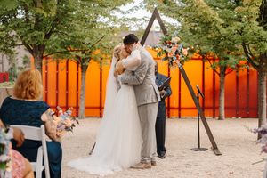 A couple dances together at an outdoor wedding venue with bright orange walls and trees in the background.