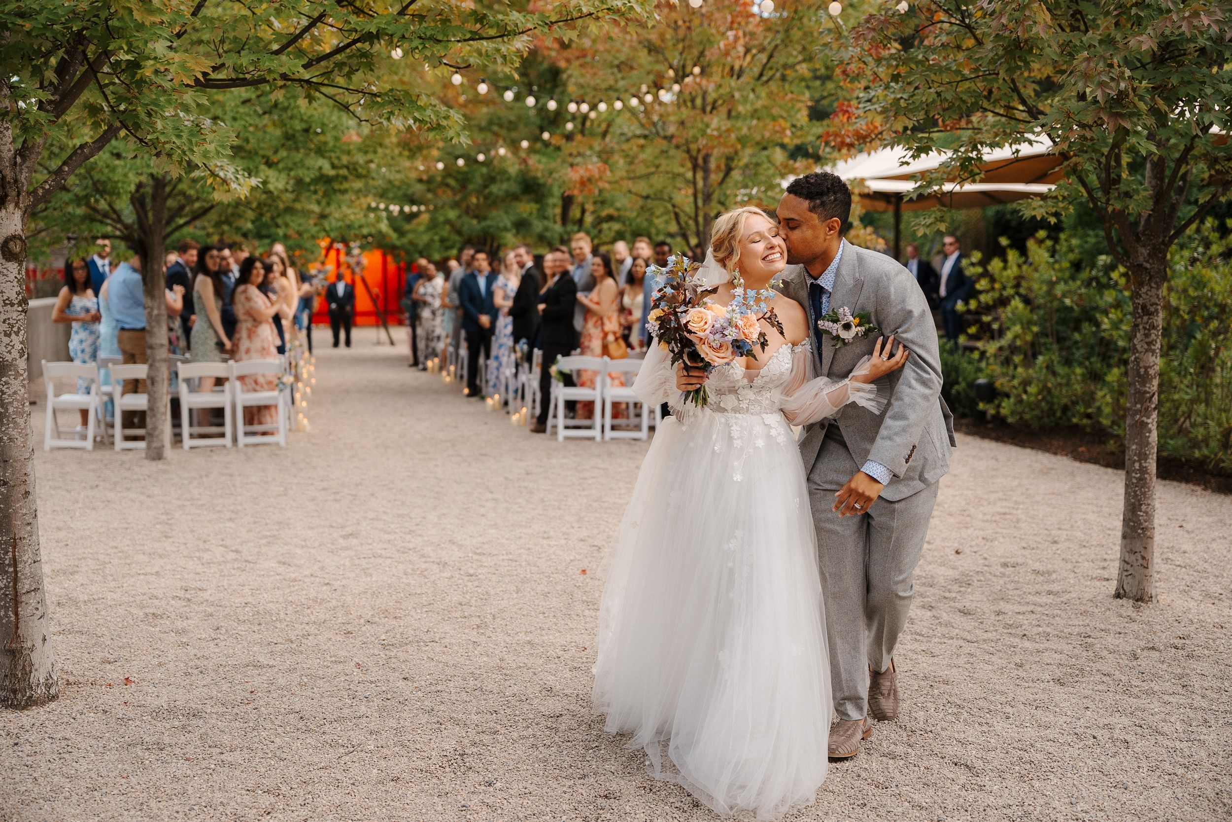 A couple shares a romantic moment at their outdoor garden wedding ceremony with guests seated on white chairs.