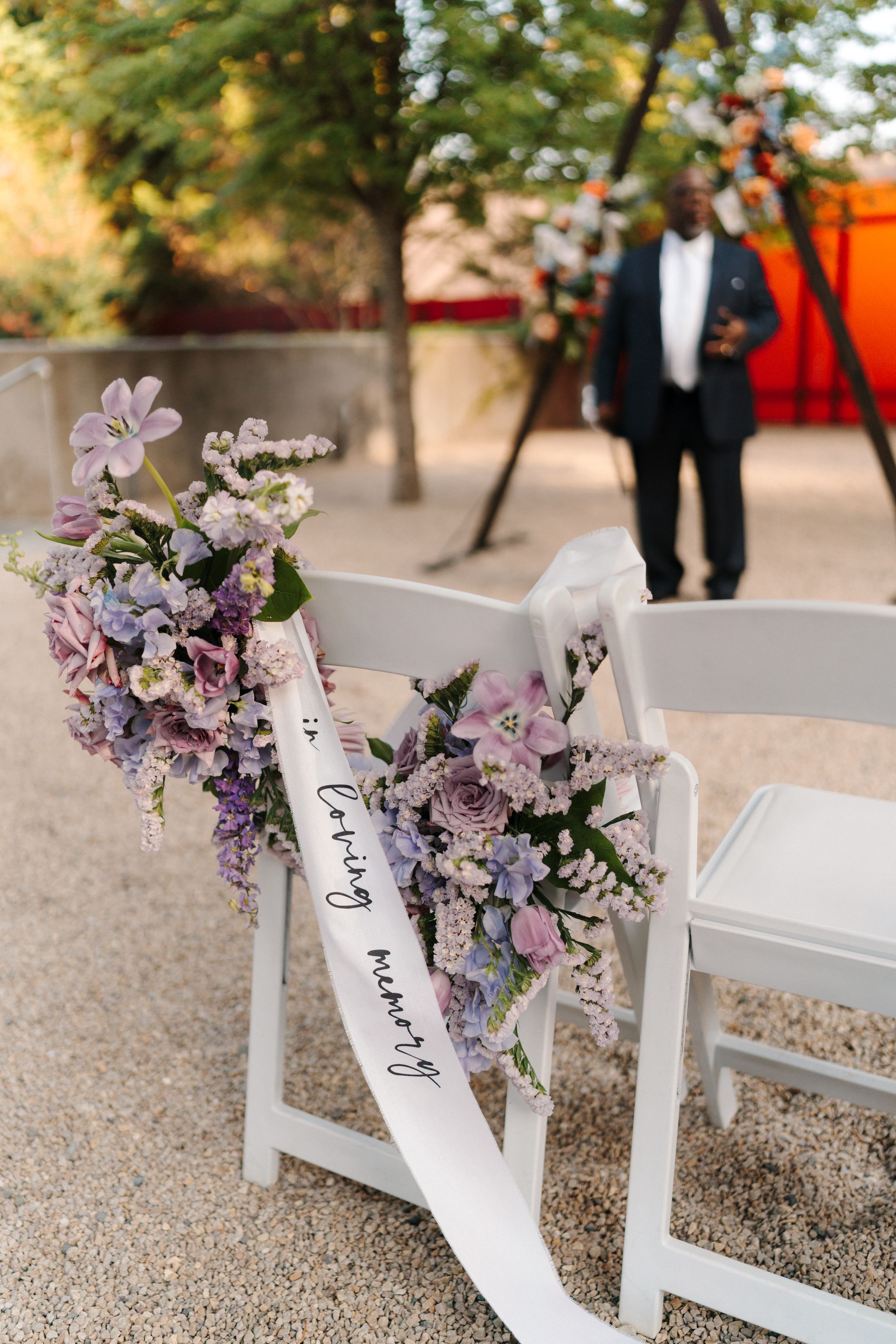 White ceremony chairs decorated with purple and lavender floral arrangements and a reserved seating ribbon at an outdoor wedding.