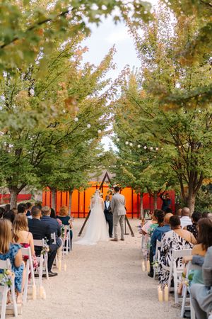 An outdoor wedding ceremony under string lights and autumn trees with a bright orange backdrop and white chairs for guests.