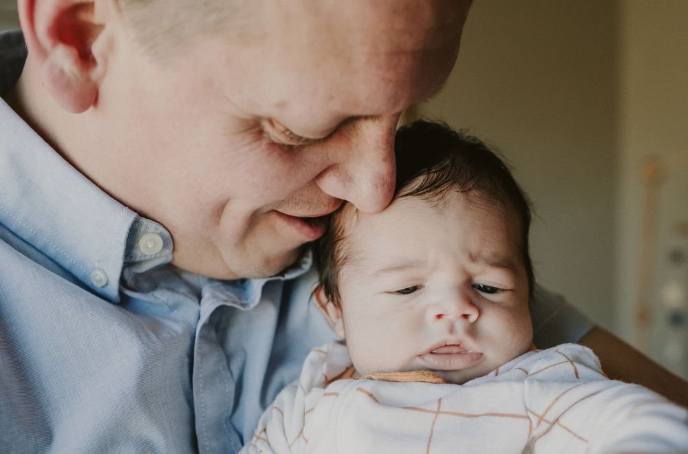 A close up of a father wearing a colored pale blue shirt holds his newborn son close to his face for newborn photos in Haverhill, MA.