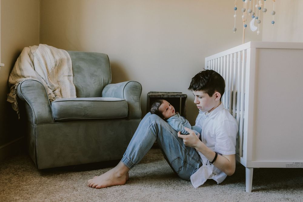 A big brother boy gazes at his newborn brother leaning against the baby's crib for newborn photos. North Shore newborn photographer.