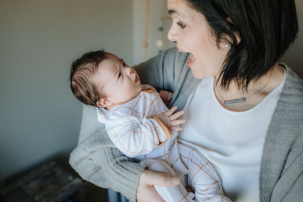 A joyful mother in a gray sweater lovingly holds and interacts with newborn wearing white in the nursery for a newborn session in MA.