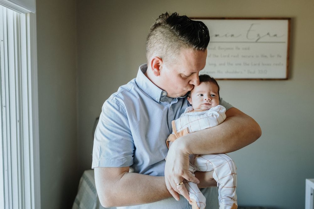 A father kisses his newborn son on the top of his head in nursery with a wooden sign in the background for a newborn photo shoot Haverhill.