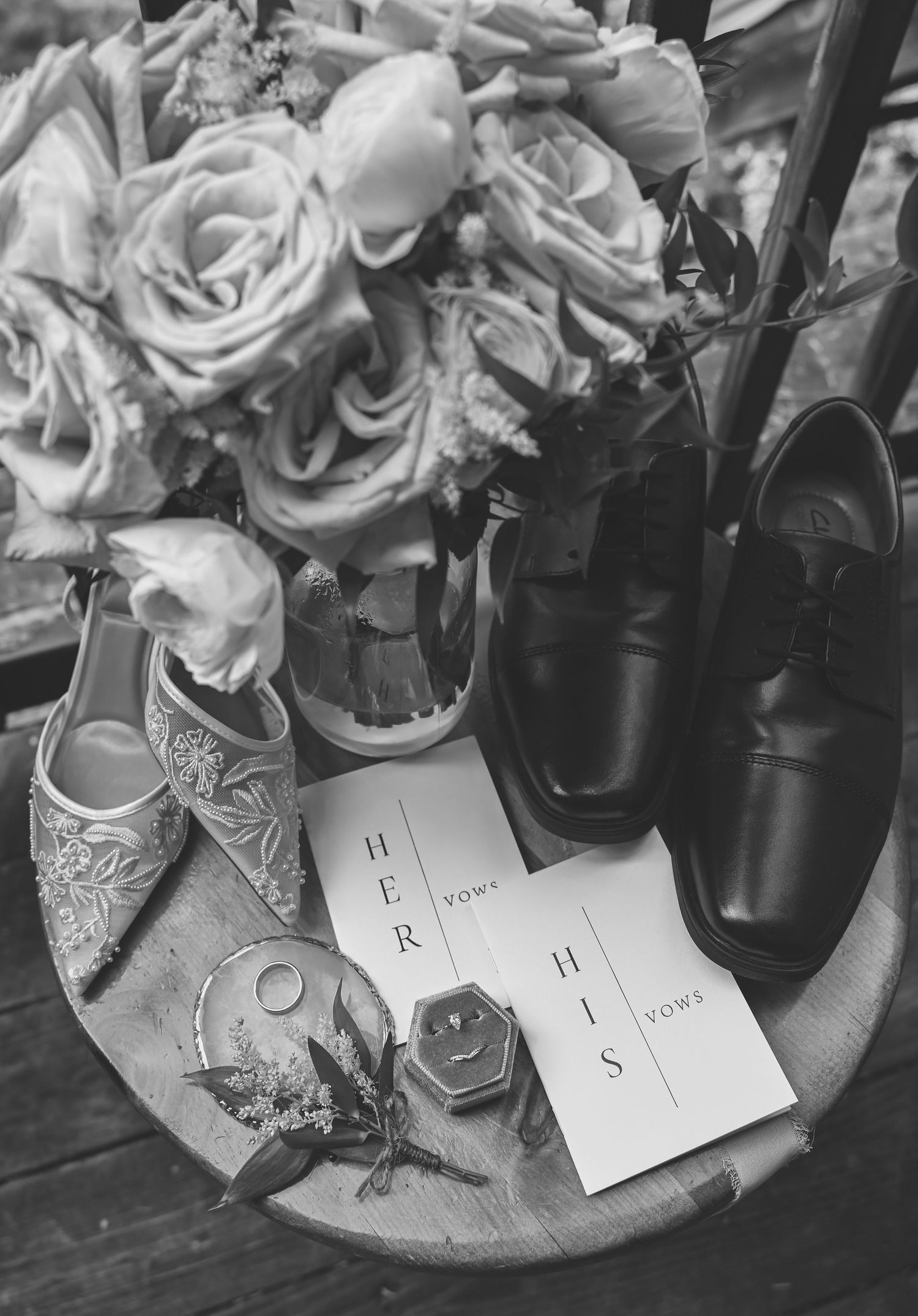 Black and white photo of wedding shoes and roses arranged with invitation cards on a round table.