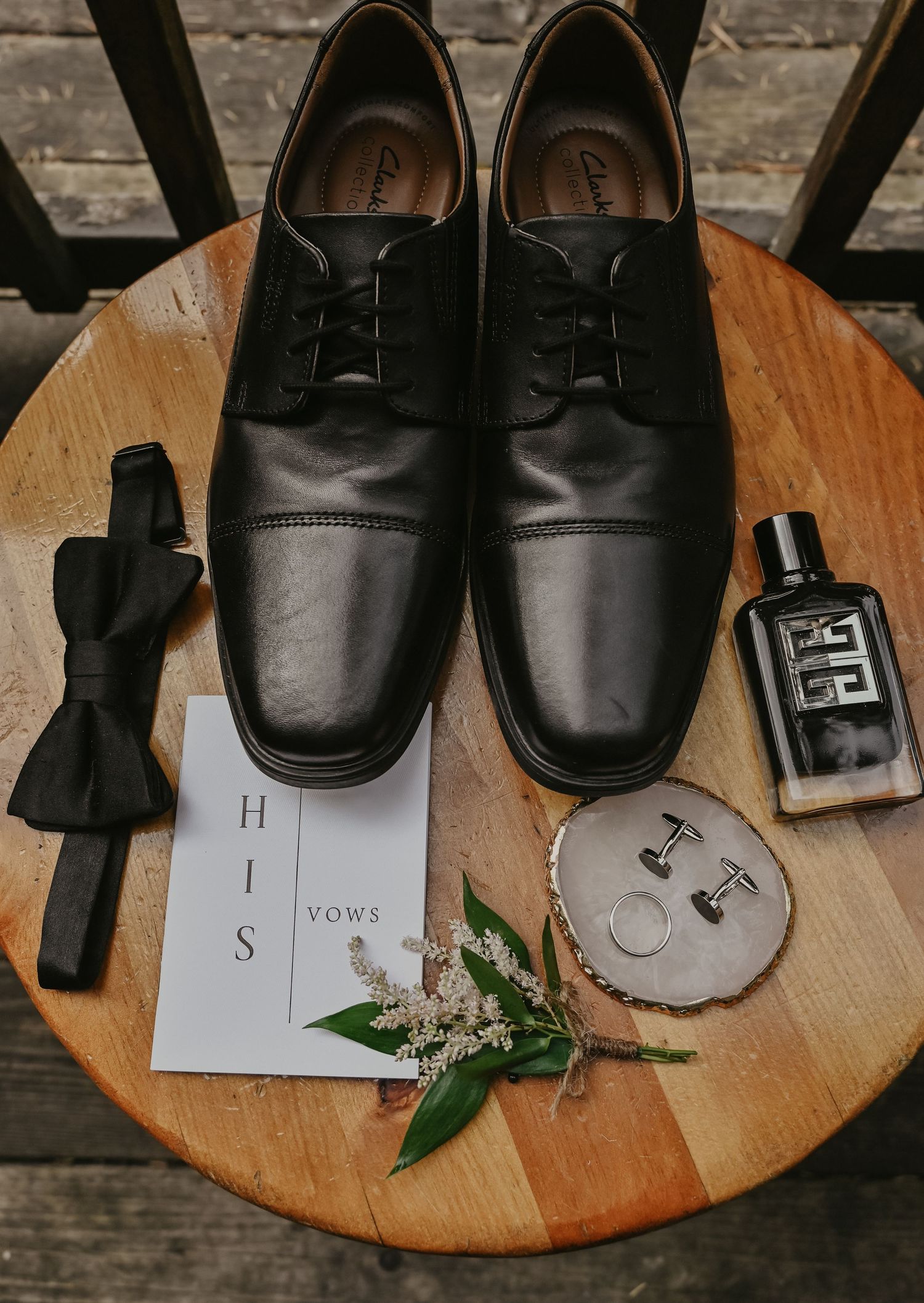 Wedding details including black dress shoes, bow tie, perfume bottle, vows card and floral sprig on wooden surface.