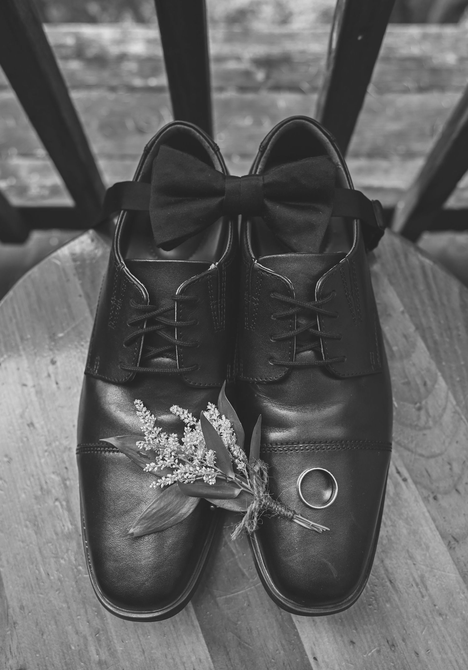 Black formal dress shoes with bow tie and delicate floral sprig arranged on wooden chair.