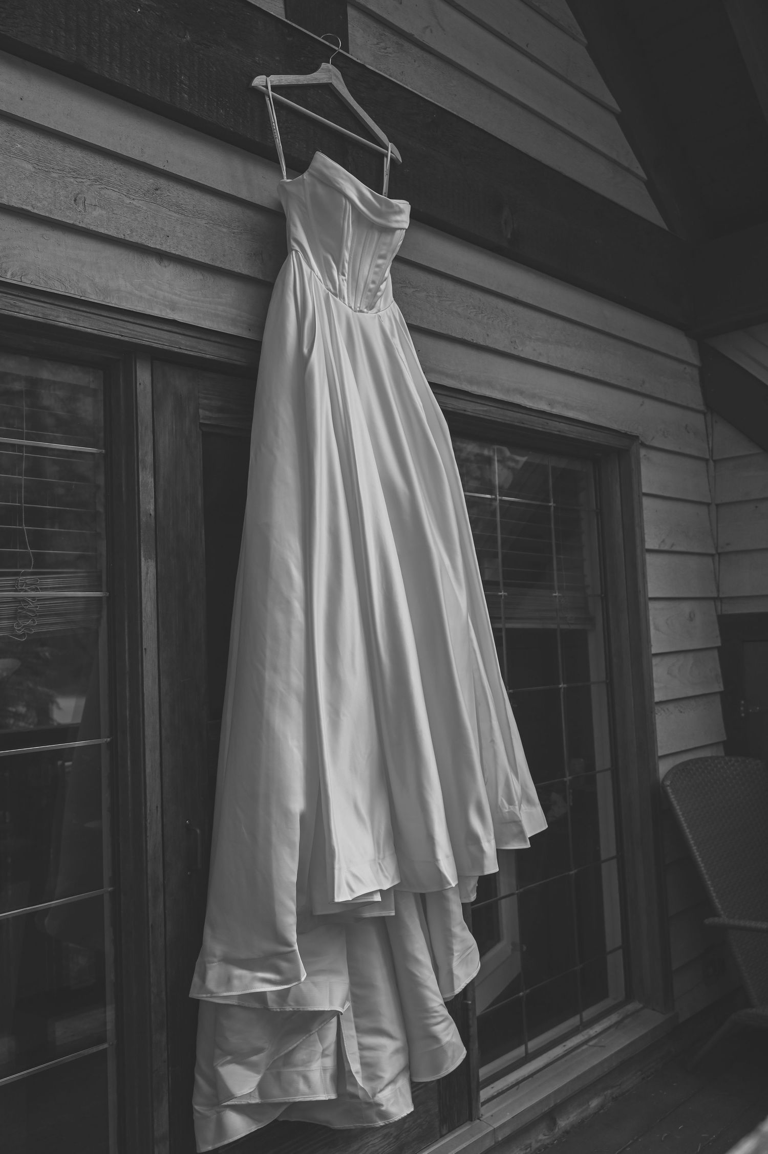A white wedding dress hangs elegantly against gray siding, photographed in black and white.