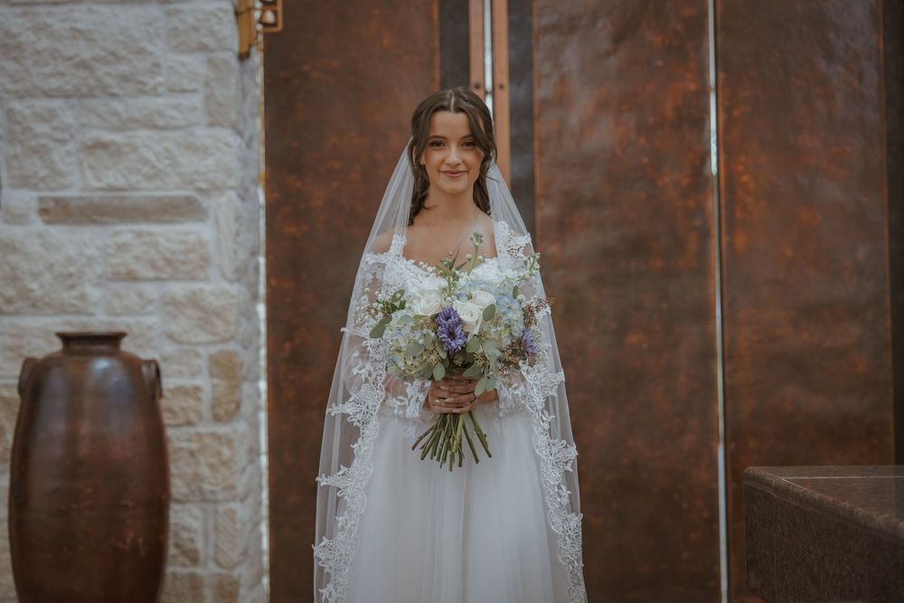 Bride in elegant white lace gown holds a natural bouquet against stone wall backdrop.