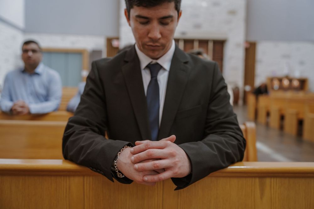Wedding guest in dark suit praying reverently in church pew.