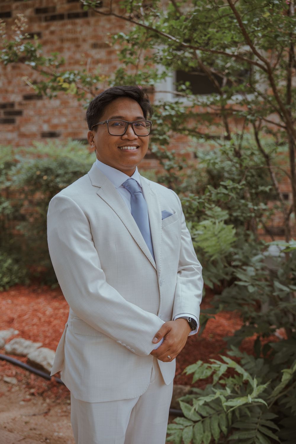 Person in white suit poses outdoors against brick wall with greenery.