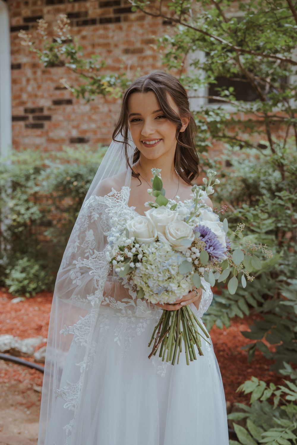 Bride holds elegant white and purple floral bouquet with lace veil.