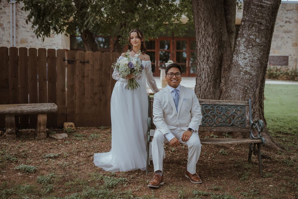 Wedding couple sits together under large tree in garden setting.