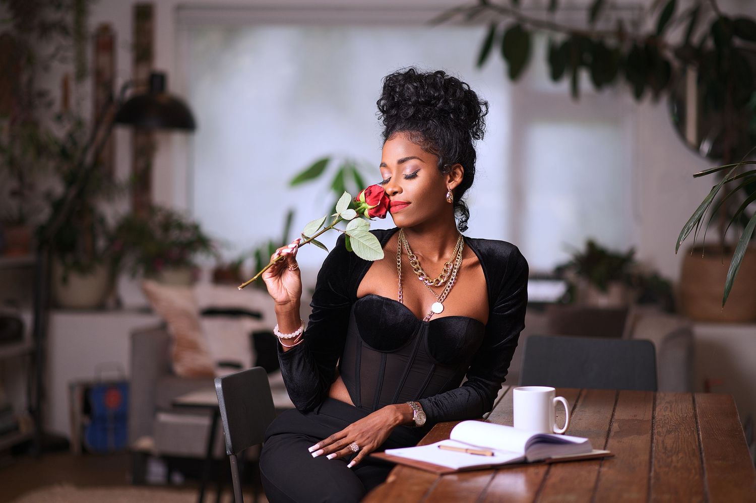 A person in a black top sits at a wooden desk writing in a notebook while surrounded by indoor plants.