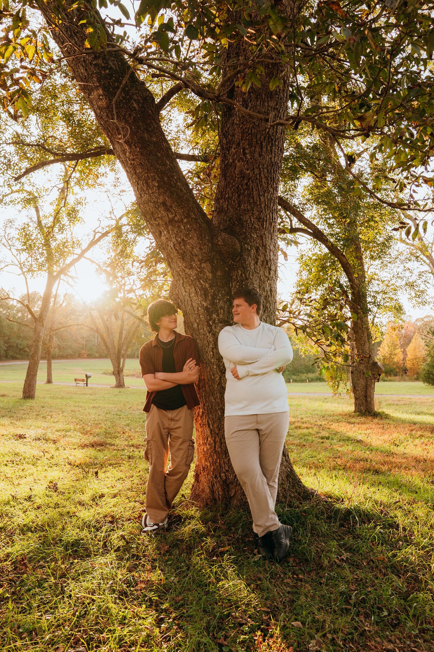 Two people share a tender moment against a tree during golden hour sunset in a grassy park setting.