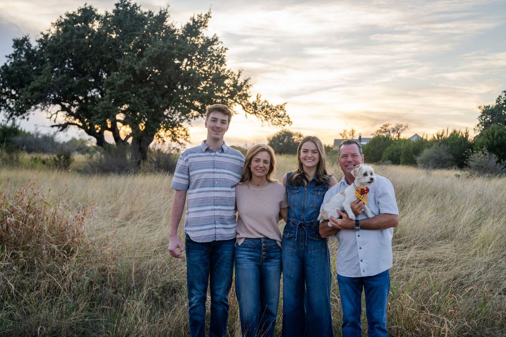 A group stands together in a grassy field with a white dog during sunset.