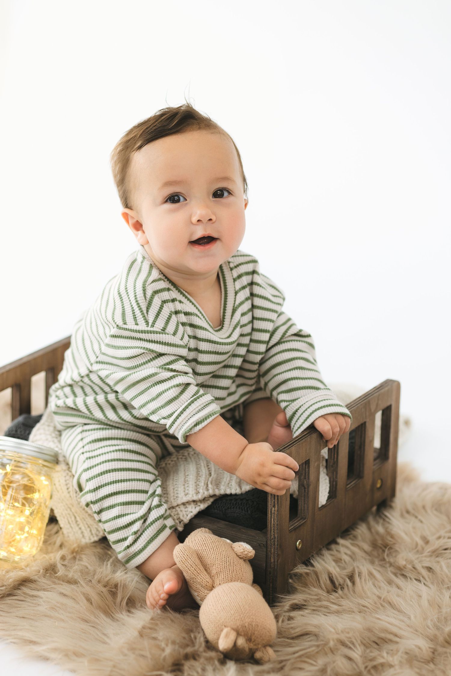 Baby in striped outfit sits on furry rug with wooden toy house and glowing fairy lights in background.